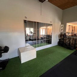 Angelika Noga Power Team - Domowa siłownia z zieloną podłogą, lustrami i sprzętem do ćwiczeń. Widoczne hantle, skrzynia do skakania i inne akcesoria. W tle bieżnia i regał z obciążeniami.