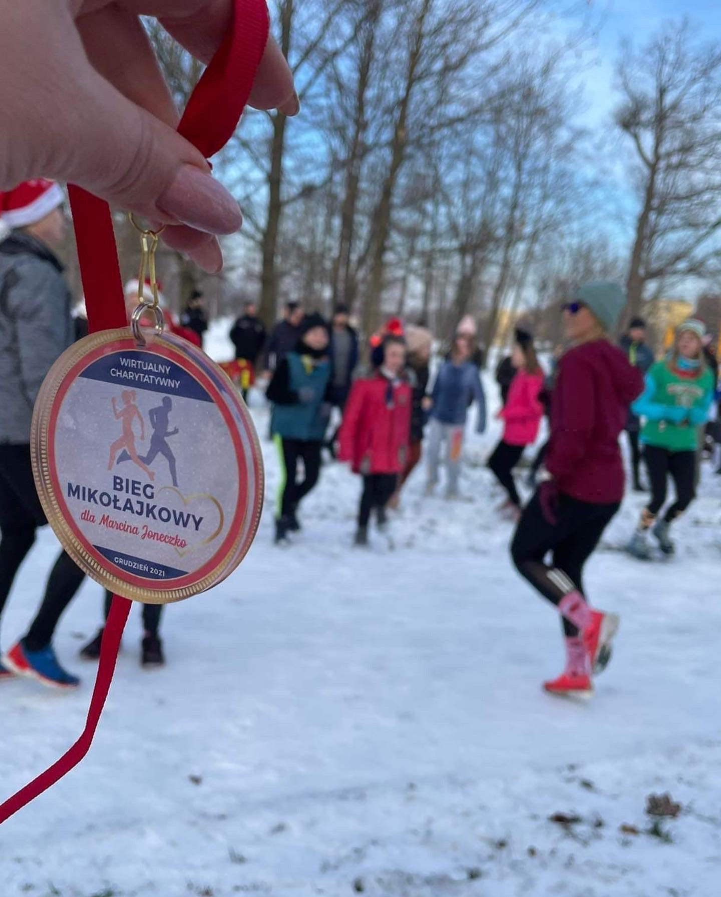 Medal 'Bieg Mikołajkowy' na czerwonej wstążce, trzymany w dłoni, z grupą biegaczy w tle na śniegu. Napis: Wirtualny Charytatywny, Grudzień 2021.