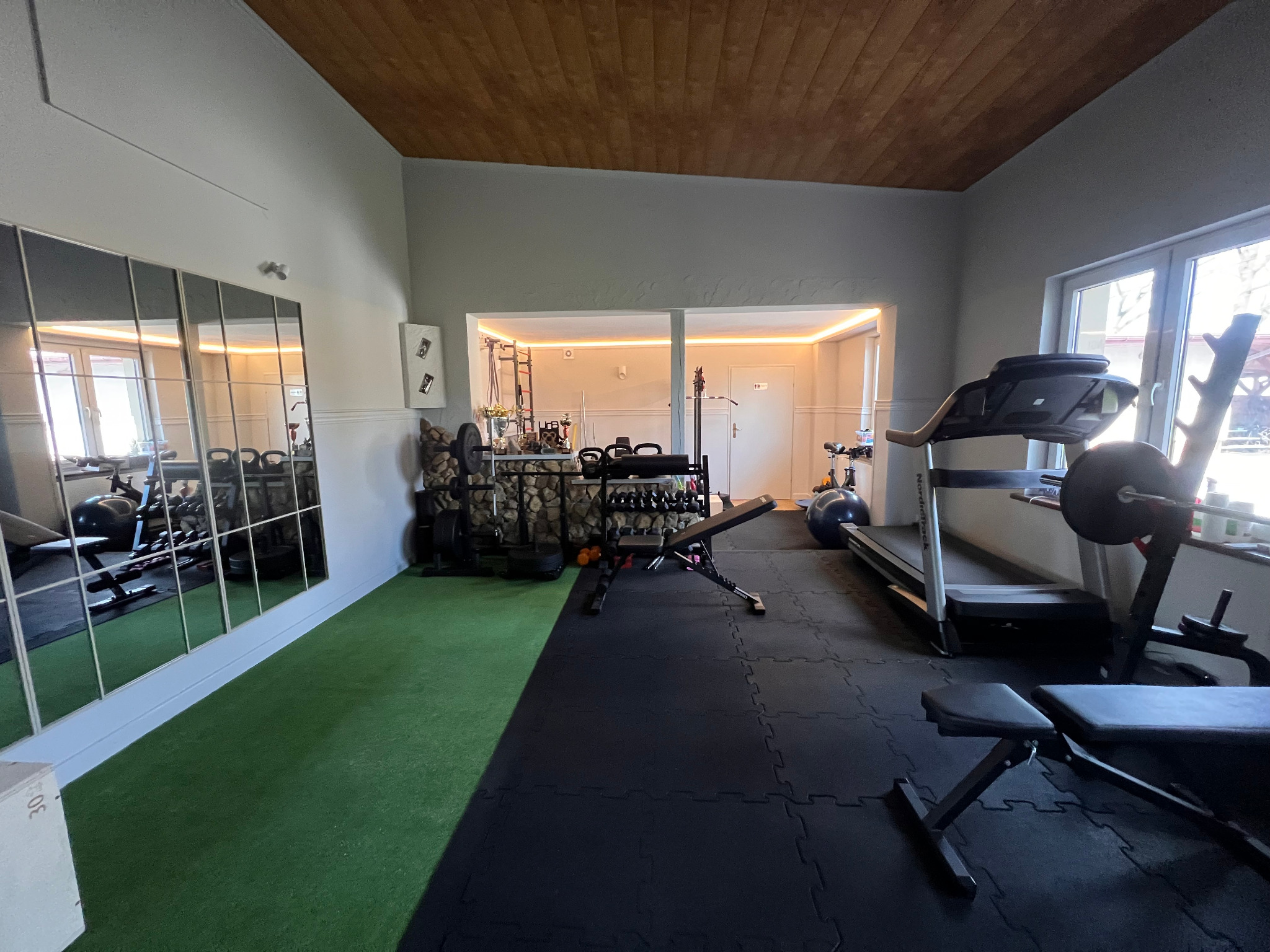 Domowa siłownia z bieżnią, ławeczką, lustrem i różnorodnym sprzętem do ćwiczeń. Podłoga wyłożona matą, sufit drewniany.