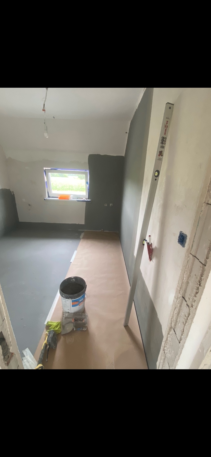 Wnętrze w trakcie prac: częściowo pomalowane ściany na szaro, przygotowane podłoże pod płytki, poziomica oparta o ścianę, wiadro z zaprawą i narzędzia glazurnicze.