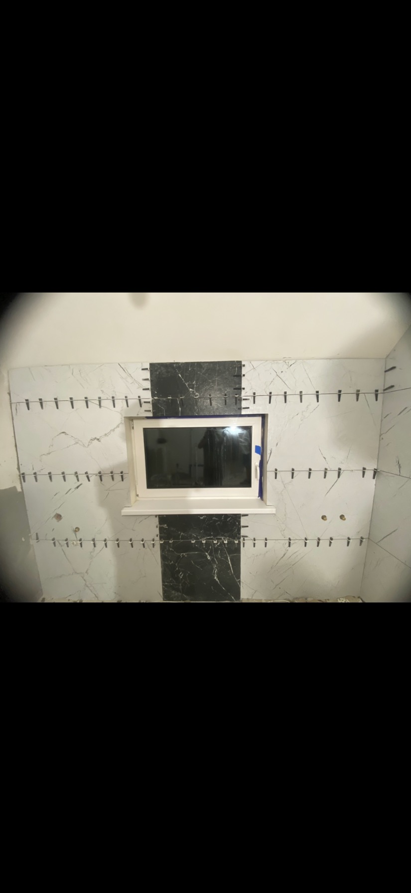 Ściana łazienki w trakcie układania płytek imitujących marmur, z czarnymi akcentami. Widoczne krzyżyki dystansowe i okno z parapetem.