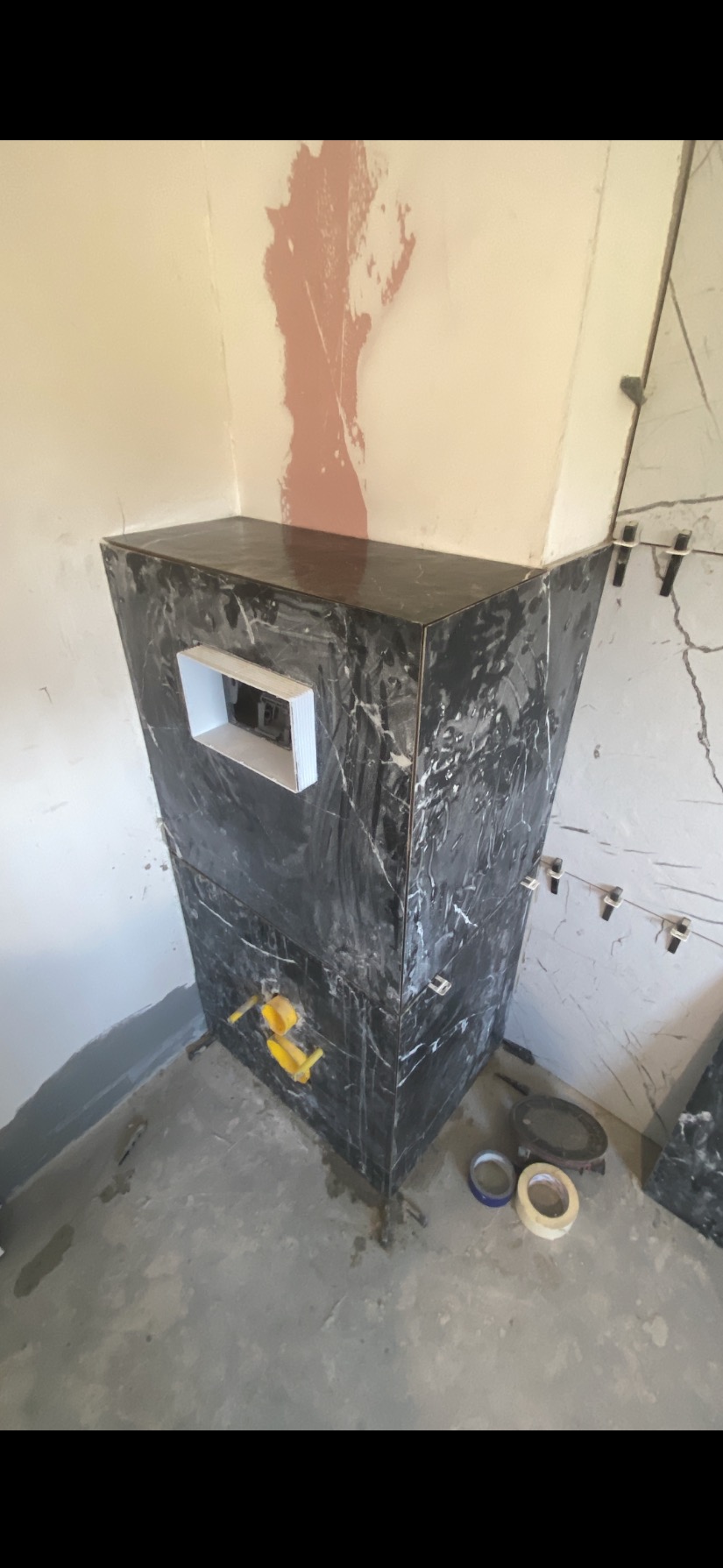 Obudowa stelaża WC z czarnego marmuru w trakcie montażu. Widoczne narzędzia glazurnicze i fragment ściany z nałożoną zaprawą. W tle płytki na ścianie.