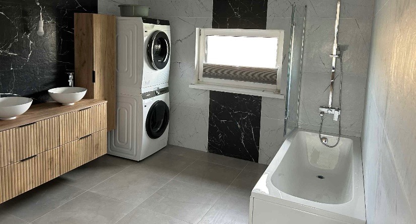 Nowoczesna łazienka z marmurowymi płytkami, drewnianą szafką z umywalkami, pralką i suszarką w kolumnie oraz białą wanną z parawanem.