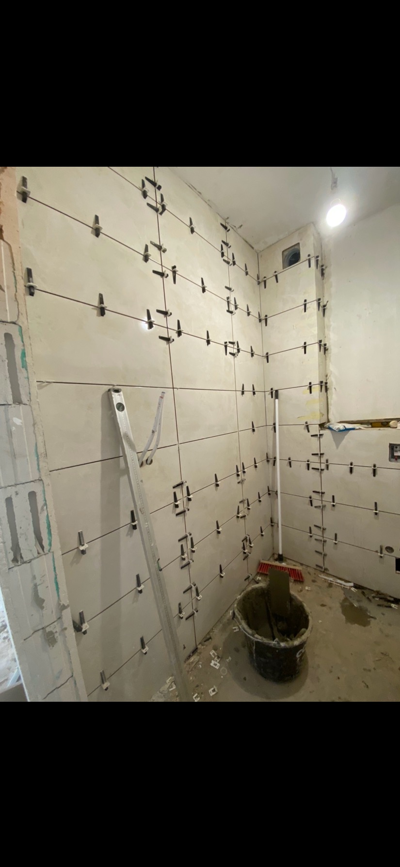 Układanie płytek w łazience: ściany z zamontowanymi krzyżykami dystansowymi, poziomica oparta o ścianę, wiadro z zaprawą. Etap prac glazurniczych.