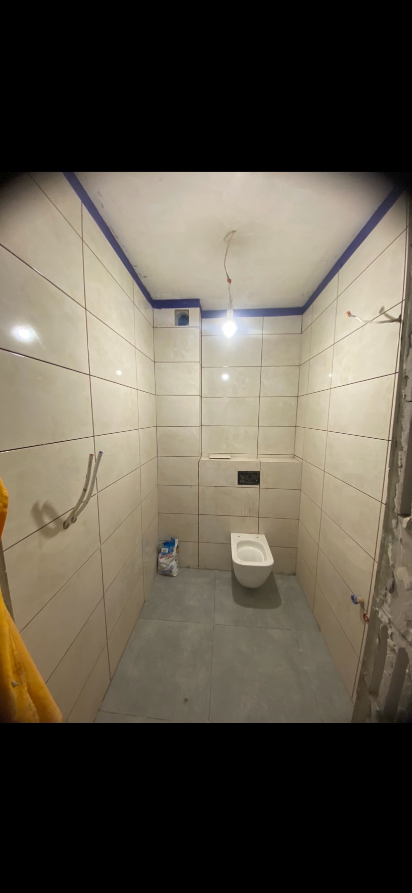 Wnętrze łazienki w trakcie wykańczania: beżowe płytki na ścianach, szare płytki na podłodze, wisząca toaleta. Widoczne ślady prac remontowych.