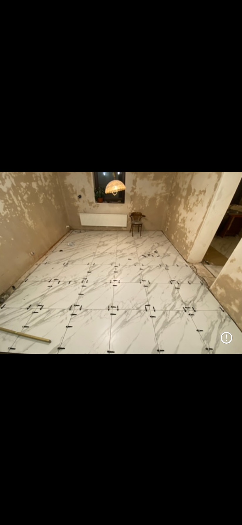 Układanie białych płytek imitujących marmur w pokoju z surowymi ścianami. Widoczne krzyżyki dystansowe. W tle okno i krzesło.