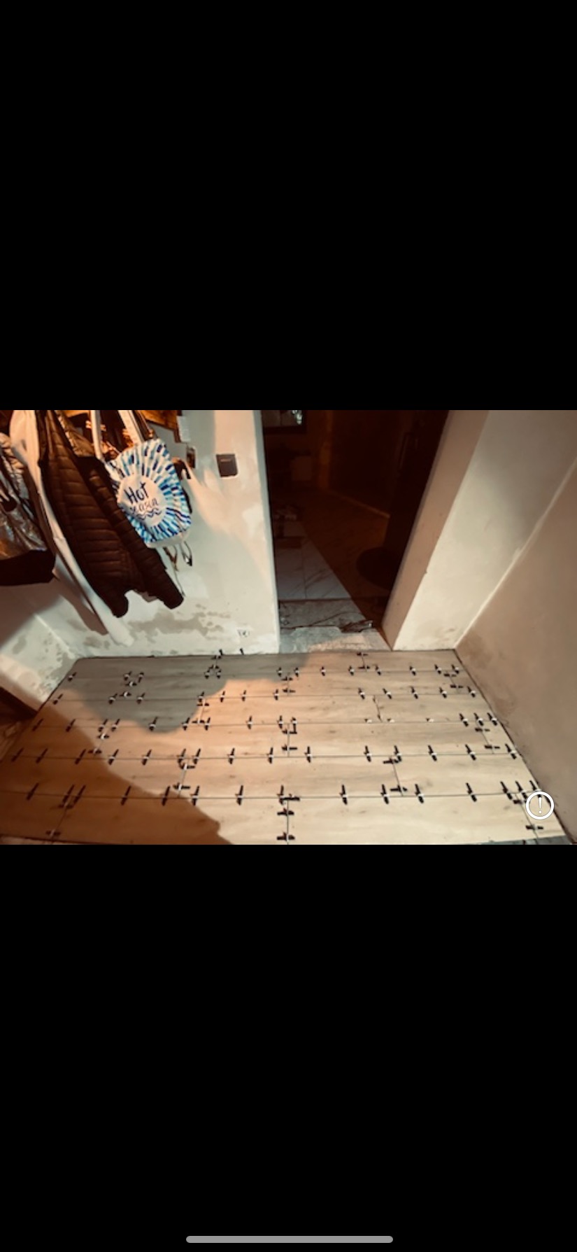 Układanie jasnych płytek podłogowych z krzyżykami dystansowymi w korytarzu. Widoczne ubrania na wieszaku i fragment sąsiedniego pomieszczenia.