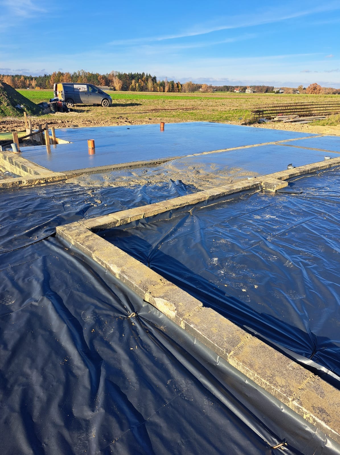 Wylana płyta fundamentowa z izolacją w postaci czarnej folii, widoczny zarys fundamentów i teren budowy z zielonym polem w tle, w oddali zaparkowany bus.