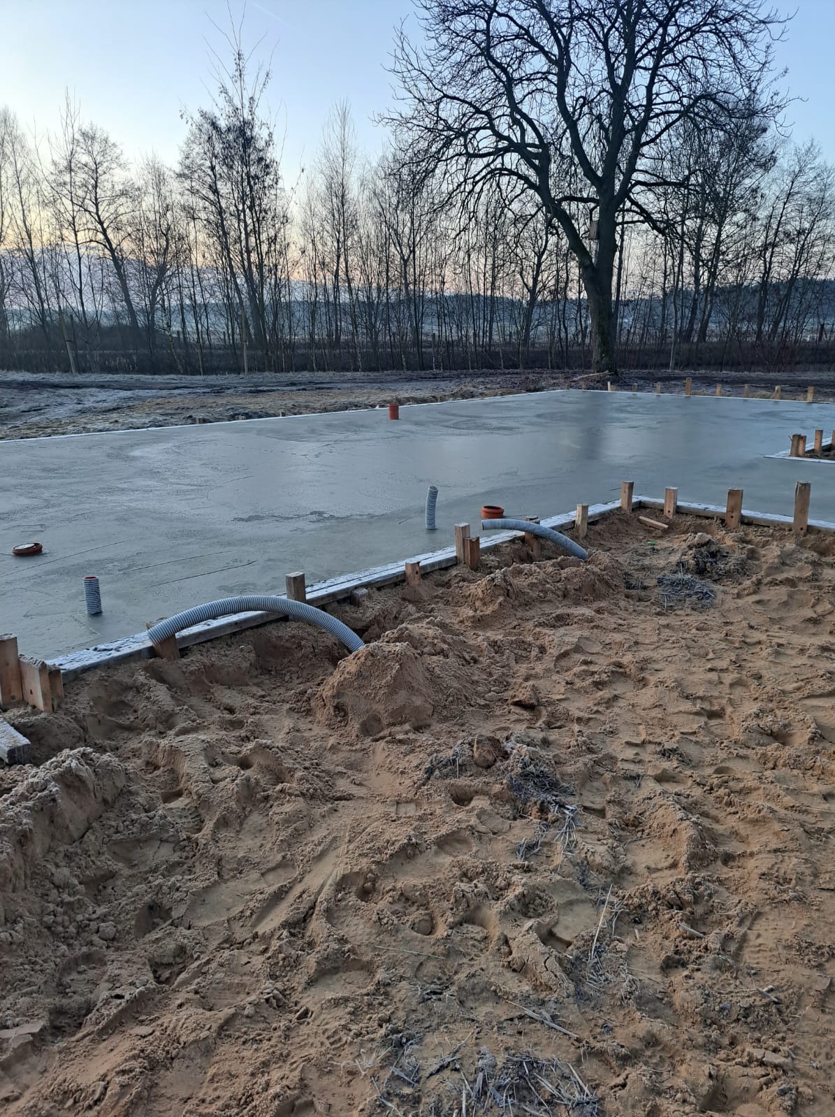 Wylana płyta fundamentowa z widocznymi elementami szalunku i instalacji w gruncie, na tle drzew bez liści i błękitnego nieba o zmierzchu.