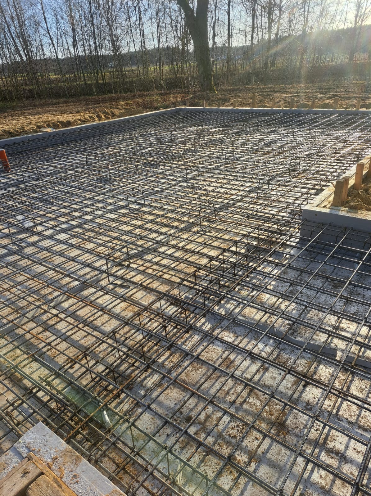 Zbrojenie płyty fundamentowej w siatkę z prętów stalowych, widok z góry, betonowe obrzeże, w tle drzewa i niebo z promieniami słońca.