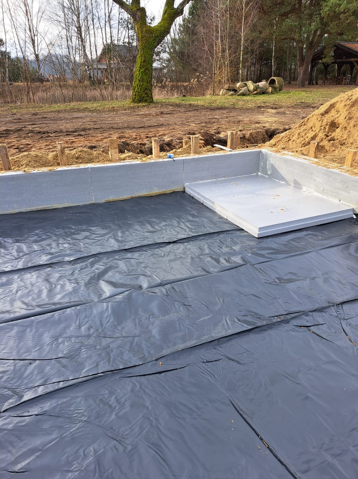 Izolacja fundamentów z folią hydroizolacyjną i szalunkiem z szarego styropianu. Widok na przygotowany teren pod budowę domu w słoneczny dzień.