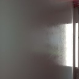 Adrian Żukowski - Świeżo pomalowana ściana w odcieniach szarości, oświetlona przez pionowy panel LED. Widoczne niedoskonałości w fakturze farby i fragment sufitu w trakcie prac.