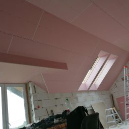 Adrian Żukowski - Montaż płyt karton-gips na poddaszu z oknami dachowymi. Widoczne narzędzia i drabina. Ściany z białego pustaka. Etap prac wykończeniowych.