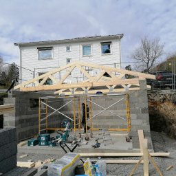 Adrian Żukowski - Budowa domu: wznoszenie ścian z bloczków betonowych i montaż drewnianej więźby dachowej. Widoczne rusztowania, narzędzia i materiały budowlane. W tle biały dom.