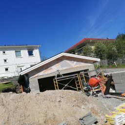 Adrian Żukowski - Budowa garażu z bloczków betonowych w trakcie realizacji. Widoczne rusztowania, mieszarka do betonu i worki z zaprawą. W tle domy jednorodzinne i błękitne niebo.