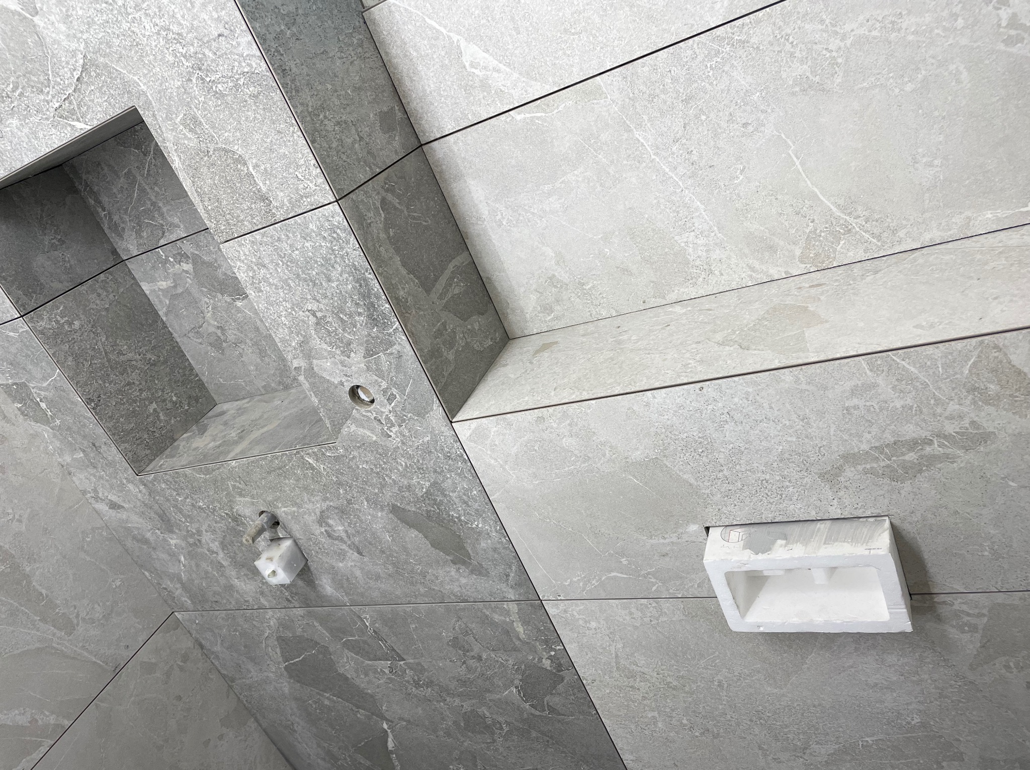 Szare, marmurowe płytki ścienne ułożone z precyzją, z wnęką i otworem na instalację. Widoczny fragment instalacji. Minimalistyczny design, nowoczesne wykończenie.