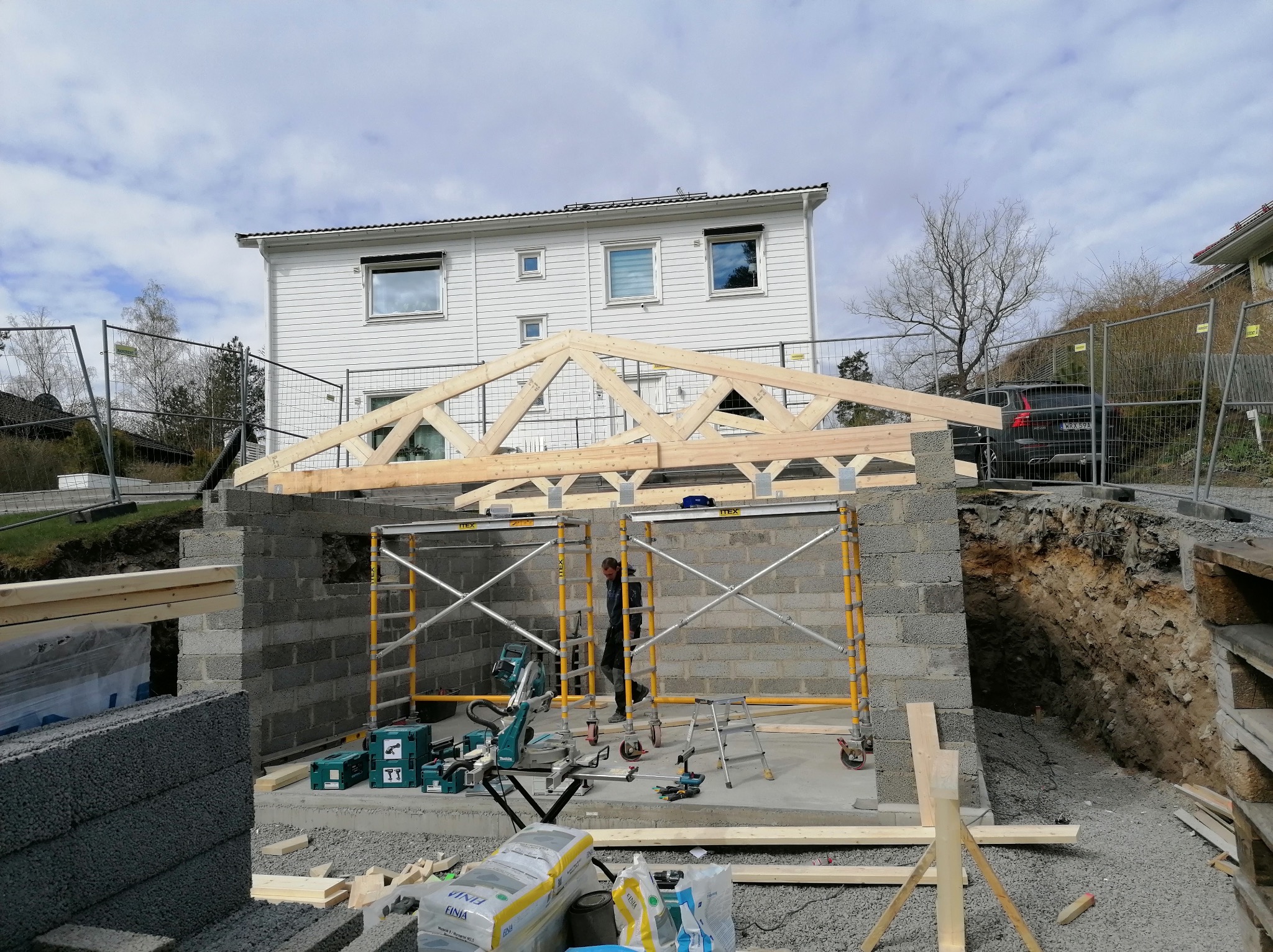 Budowa domu: wznoszenie ścian z bloczków betonowych i montaż drewnianej więźby dachowej. Widoczne rusztowania, narzędzia i materiały budowlane. W tle biały dom.