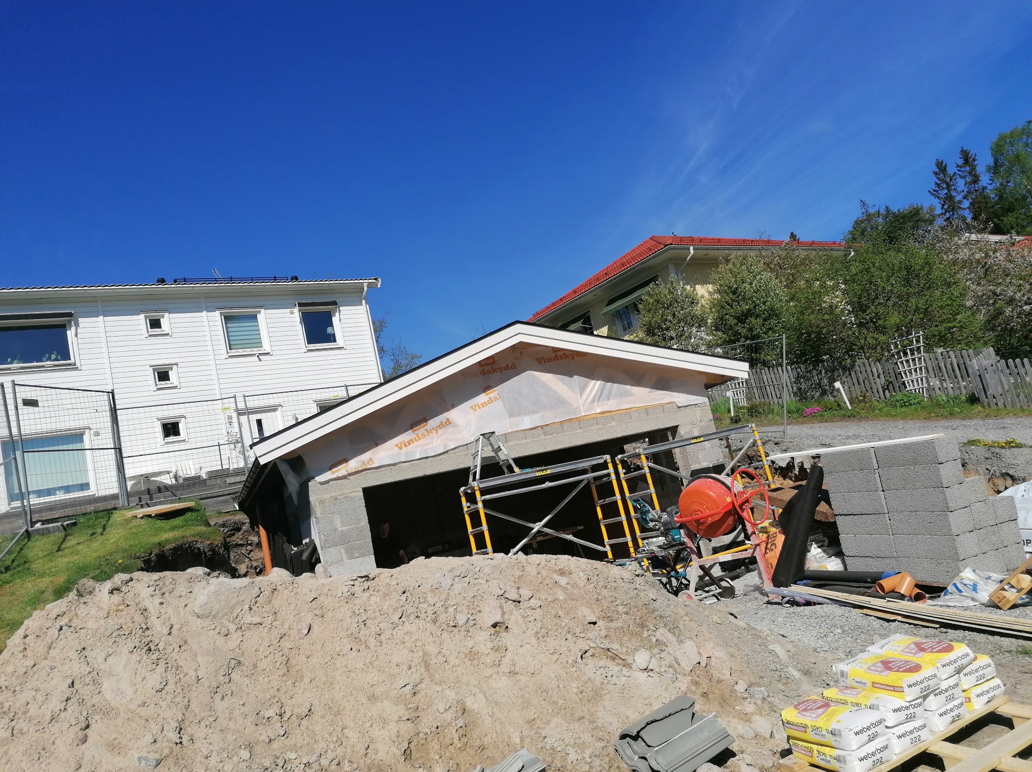 Budowa garażu z bloczków betonowych w trakcie realizacji. Widoczne rusztowania, mieszarka do betonu i worki z zaprawą. W tle domy jednorodzinne i błękitne niebo.
