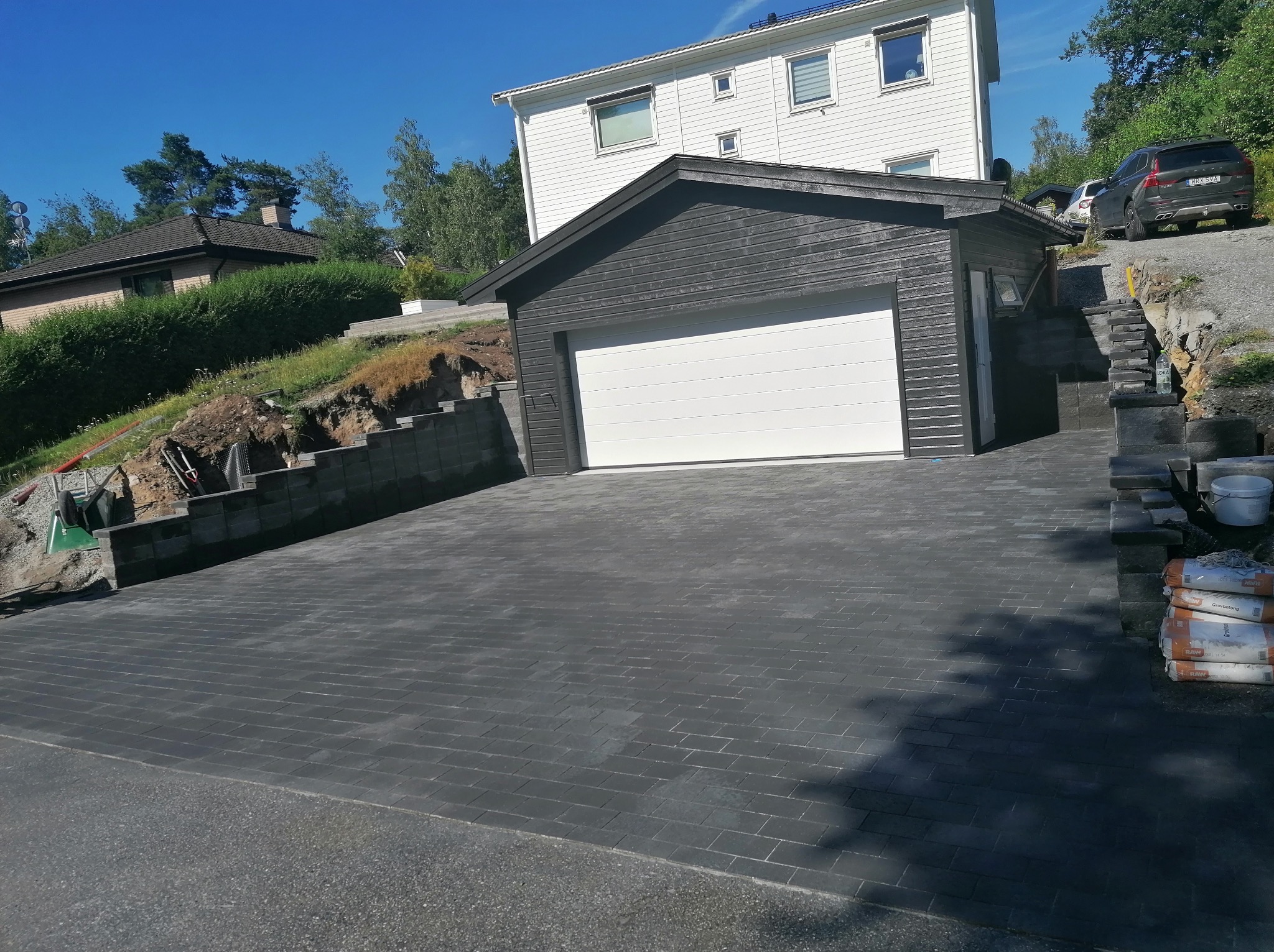 Ułożona, grafitowa kostka brukowa na podjeździe do garażu z białą bramą, na tle domu z białymi ścianami i błękitnego nieba. Widoczne worki z cementem.