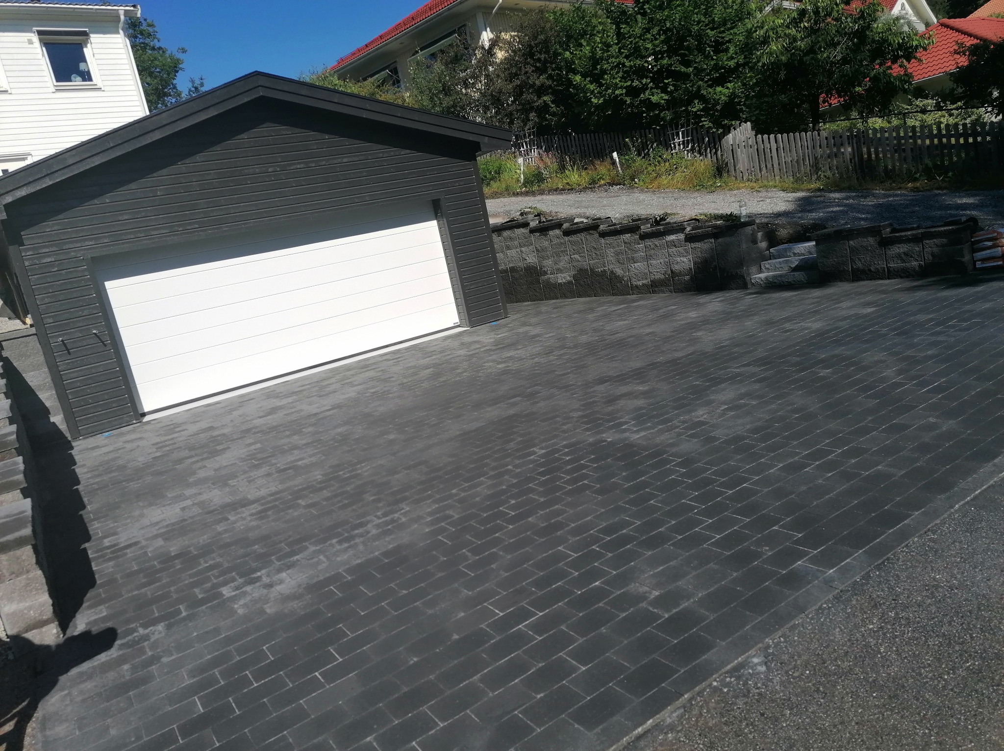 Nowa, ciemnoszara kostka brukowa przed garażem z białą bramą, otoczona zielenią i elementami architektury ogrodowej. Ujęcie z niskiej perspektywy.
