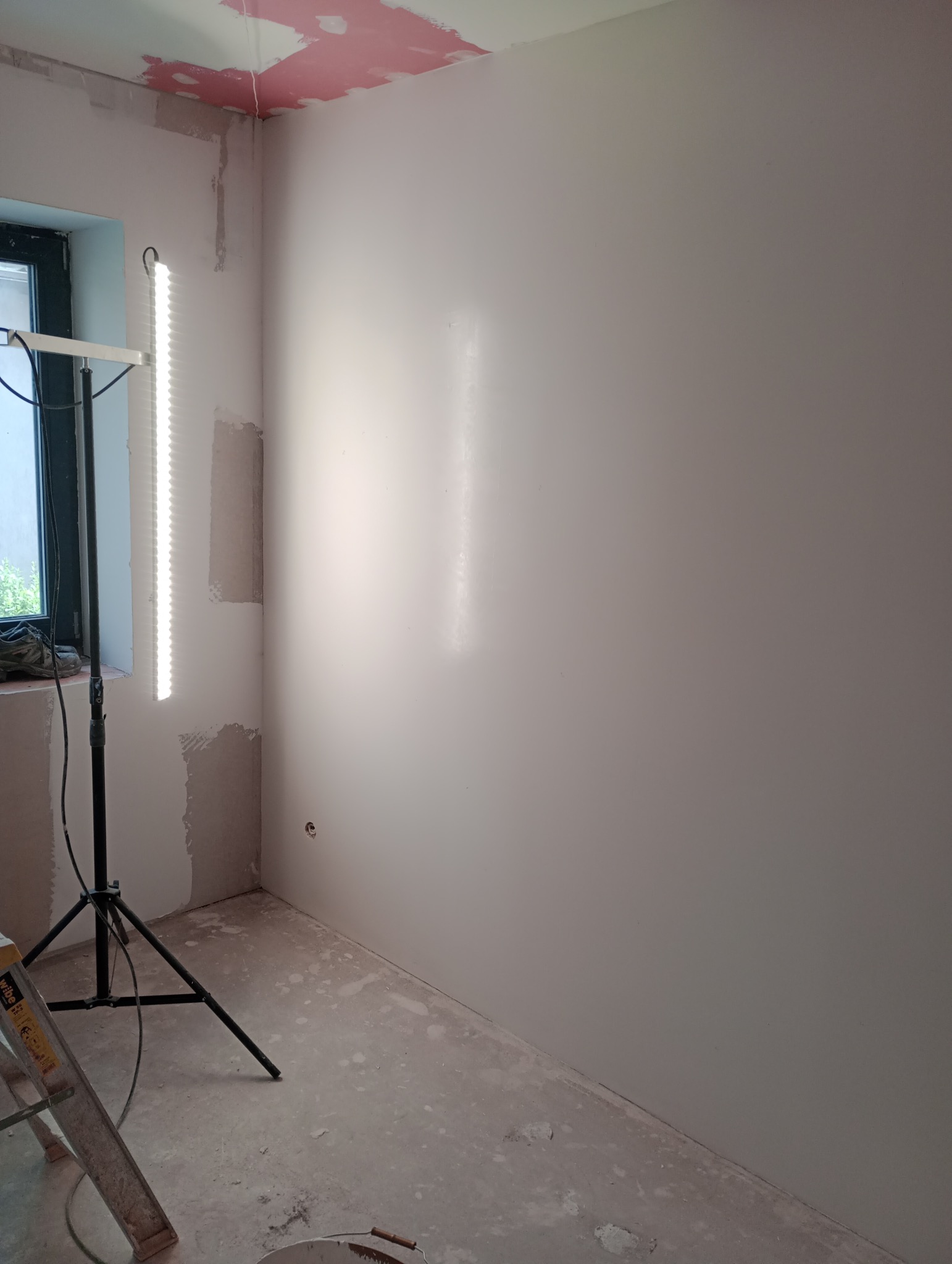 Pokój w trakcie szpachlowania: gładkie ściany, ślady gipsu na suficie, lampa budowlana i drabina. Widoczne niedoskonałości podłoża przed malowaniem.
