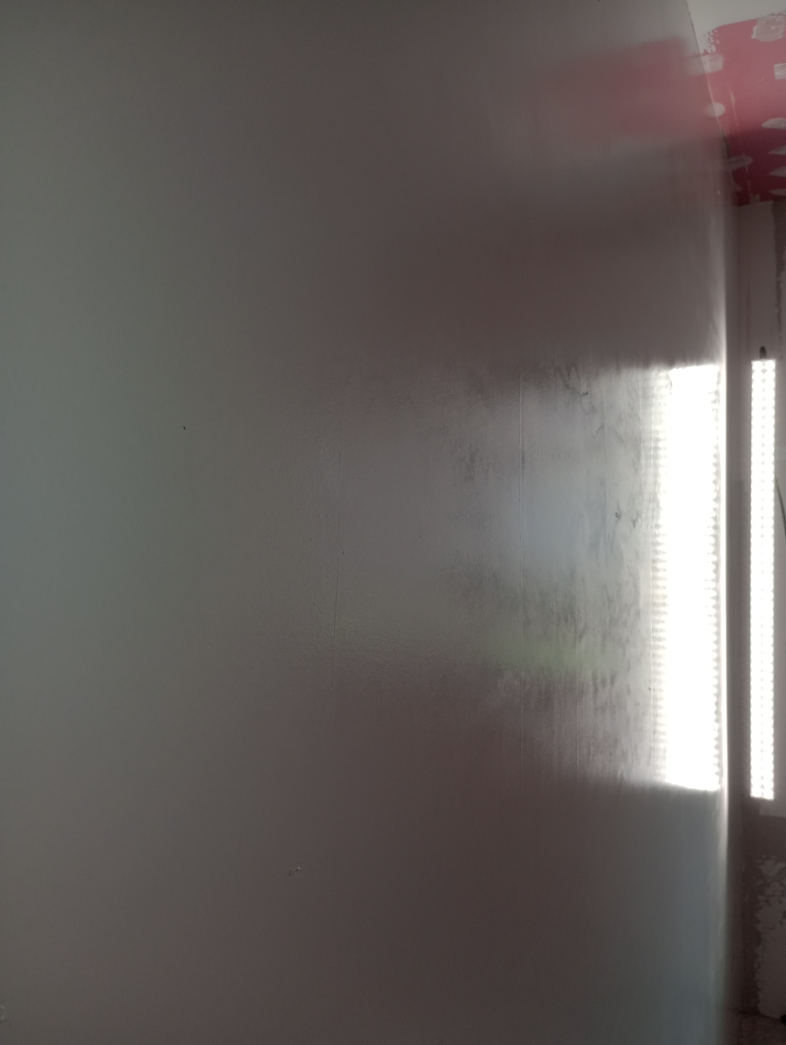 Gładka, szara ściana po szpachlowaniu, oświetlona bocznym światłem lampy LED. Widoczne delikatne nierówności i faktura powierzchni. Fragment sufitu z resztkami taśmy malarskiej.