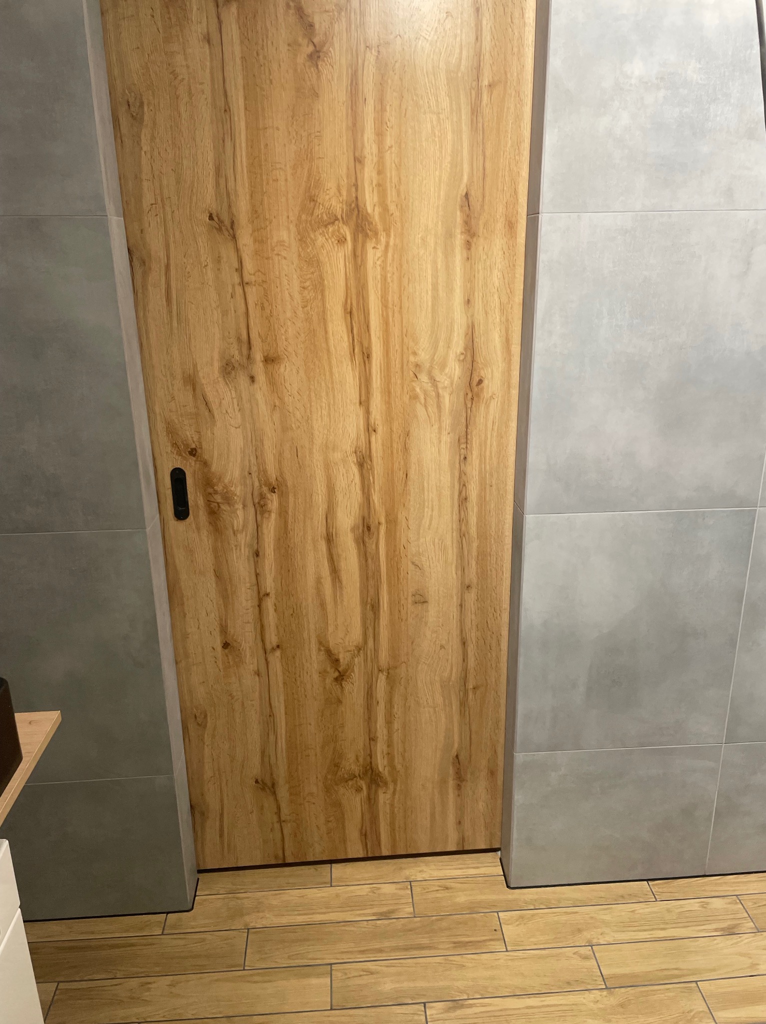 Drewniane drzwi przesuwne z czarnym uchwytem, wkomponowane w szarą, kwadratową glazurę ścienną i podłogę z paneli imitujących drewno. Detal wykończenia wnętrza.