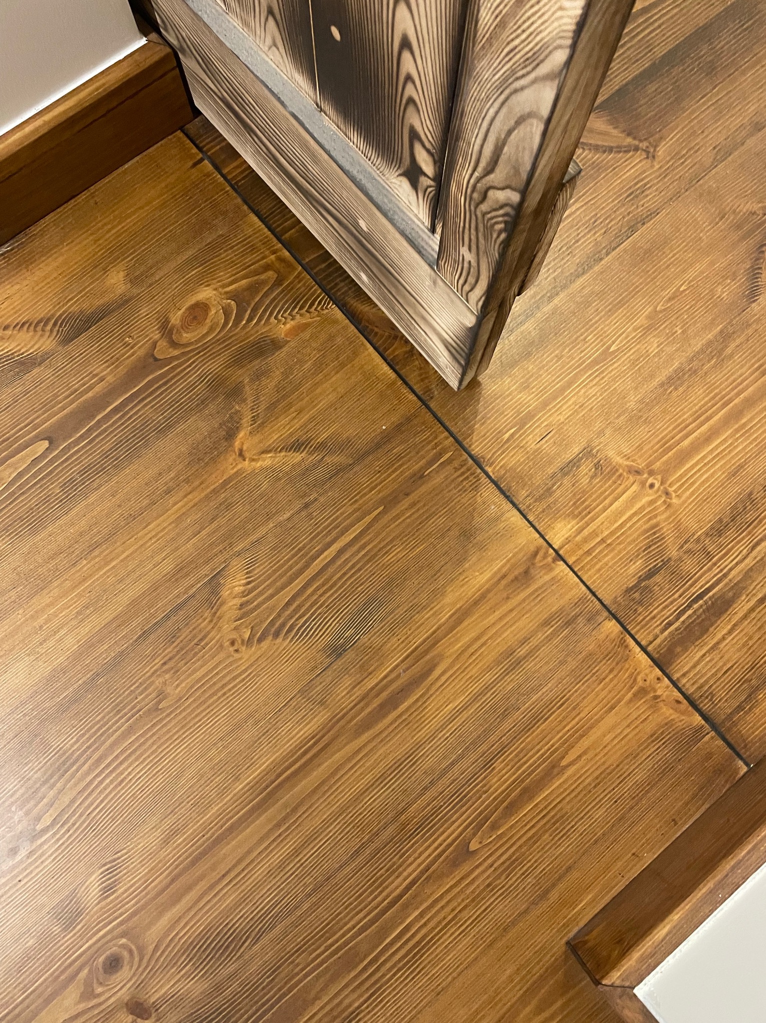 Lakierowana podłoga z desek o ciepłym, miodowym odcieniu, z widocznym usłojeniem drewna. Fragment drzwi z ciemnego, opalizowanego drewna w rogu kadru.