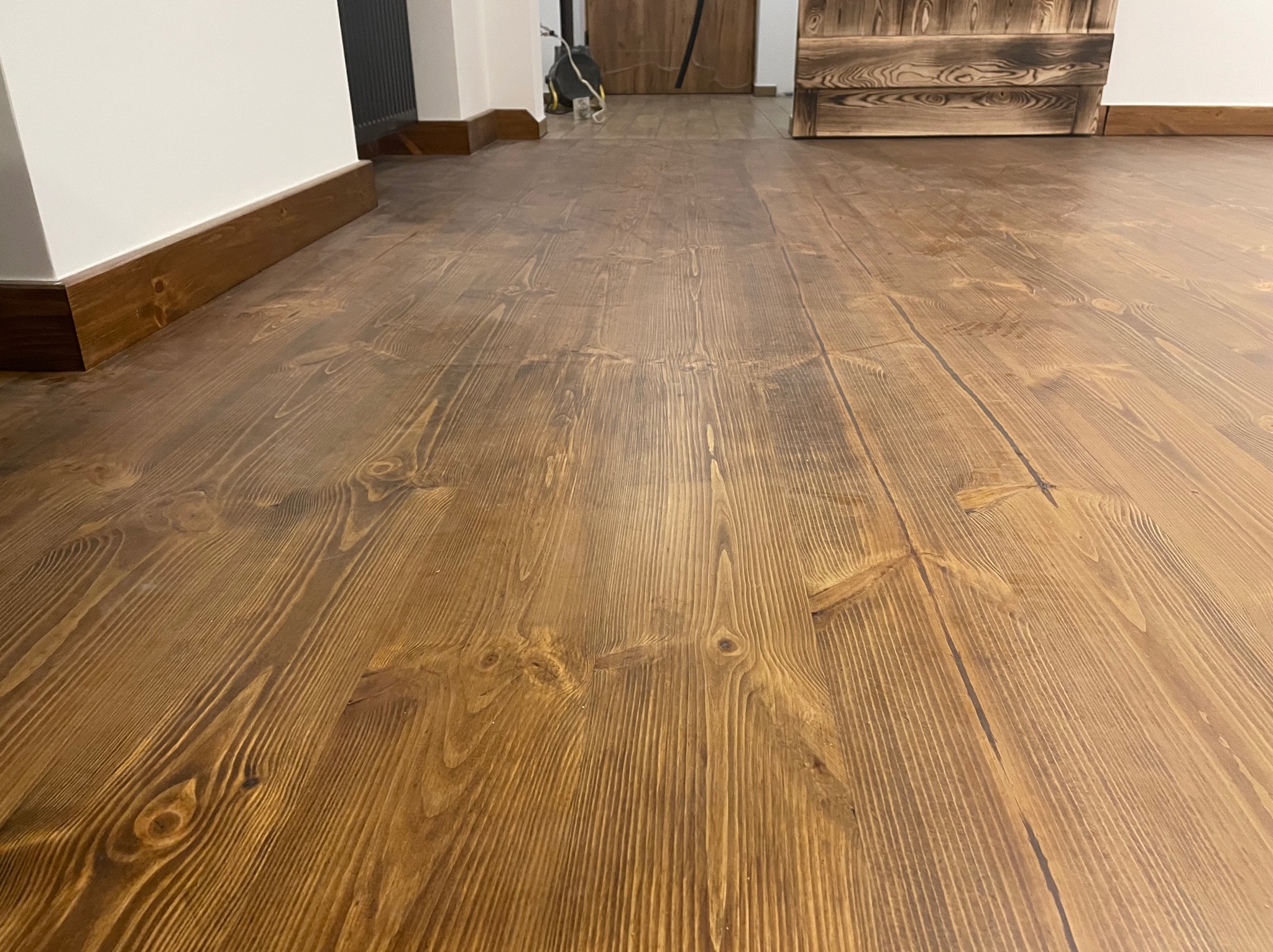 Lakierowana podłoga z desek o ciepłym, brązowym odcieniu, z widocznymi słojami drewna. Listwy przypodłogowe w kolorze podłogi. W tle drzwi i fragment grzejnika.