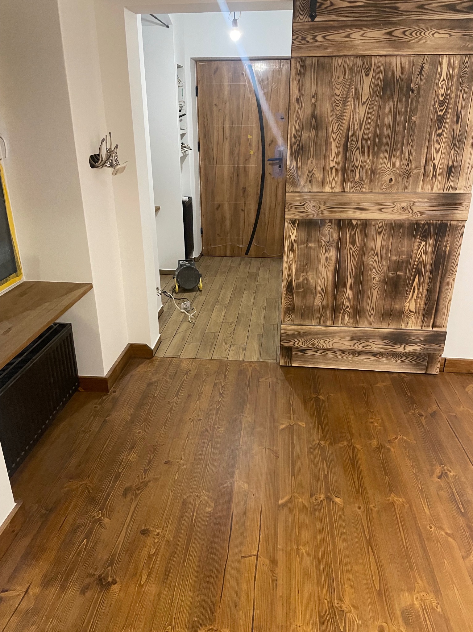 Rustykalne, drewniane drzwi przesuwne z palonym wzorem, widoczne na tle drewnianej podłogi w ciepłym odcieniu. W tle widoczny fragment przedpokoju.