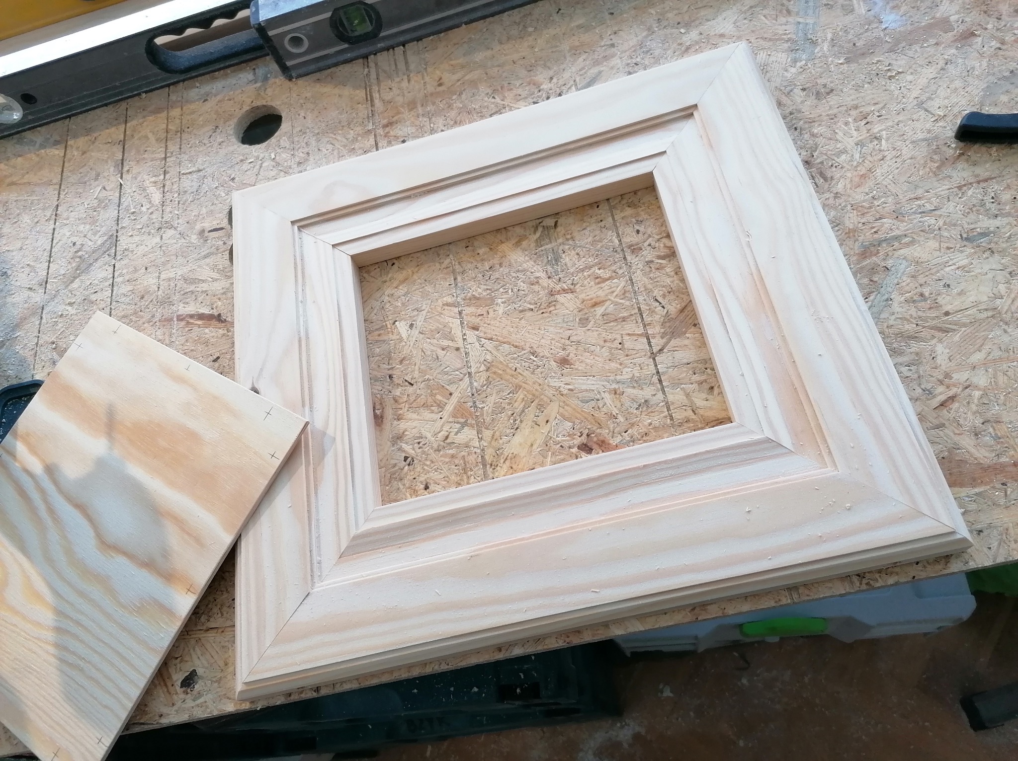 Drewniana rama z listwami dekoracyjnymi, leżąca na płycie OSB. Obok leży deska z zaznaczonymi punktami. Widoczna poziomica i narzędzia stolarskie.