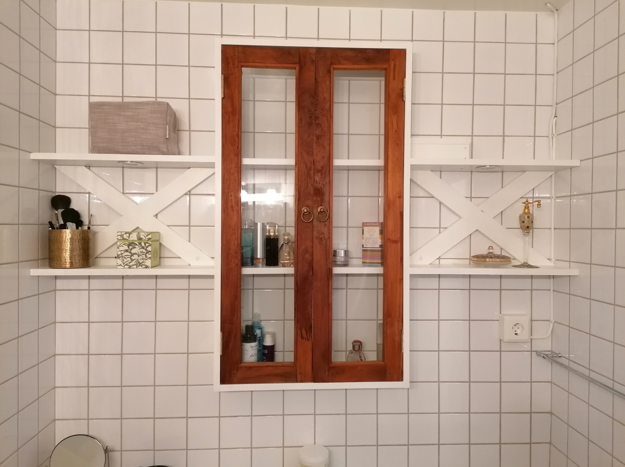 Białe kafelki w łazience z drewnianą szafką z przeszklonymi drzwiczkami i białymi półkami z dekoracjami. Widoczny fragment lustra i gniazdko elektryczne.