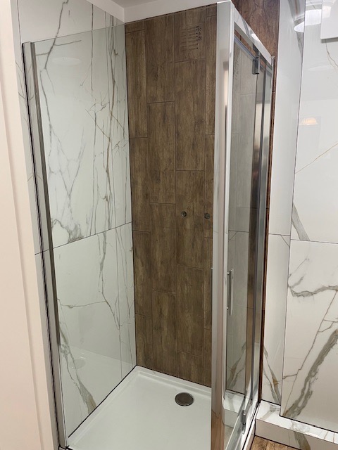 Nowoczesna kabina prysznicowa z przesuwnymi drzwiami, wykończona płytkami imitującymi marmur i drewno, z widocznym odpływem liniowym.