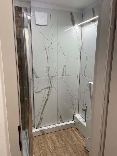 Wykończona kabina prysznicowa z białymi płytkami imitującymi marmur i drewnopodobną podłogą. Widoczny wentylator i oświetlenie LED.