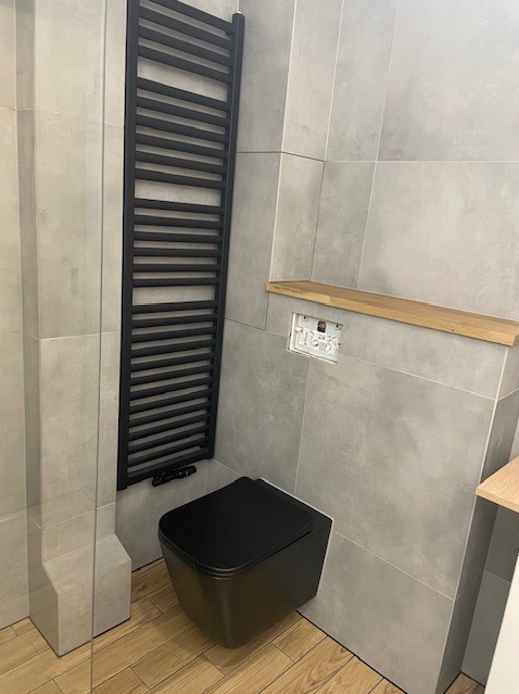 Nowoczesna łazienka z szarymi płytkami, czarnym grzejnikiem drabinkowym i podwieszaną toaletą. Drewniane półki dodają ciepła minimalistycznemu wnętrzu.