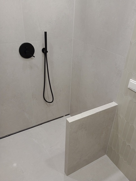 Nowoczesna kabina prysznicowa z czarną armaturą i odpływem liniowym, wykończona jasnymi płytkami w odcieniach szarości. Minimalistyczny design z akcentem na detale.