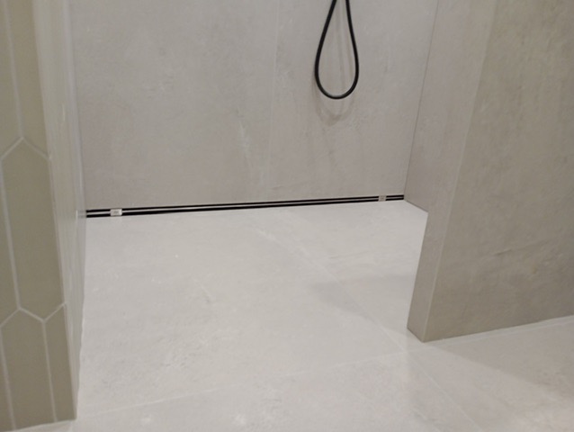 Wykończona kabina prysznicowa z odpływem liniowym i jasnymi płytkami. Widoczne detale wykończenia ścian i podłogi, minimalistyczny design.