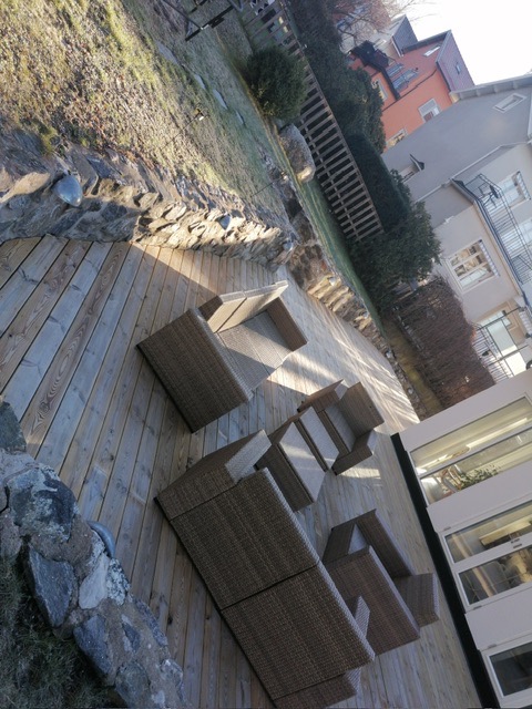 Drewniany taras z rattanowymi meblami ogrodowymi, otoczony kamiennym murkiem i zielenią. Ustawiony pod kątem, z widokiem na budynek i ogród.