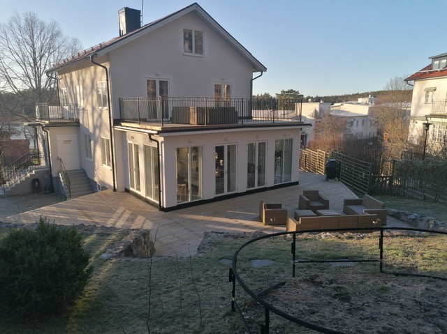 Biały dom z balkonami i przeszkloną werandą, otoczony drewnianym tarasem i ogrodem. Widok z góry, w tle zabudowa jednorodzinna i drzewa.