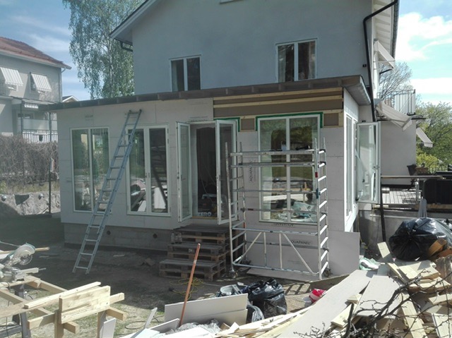 Rozbudowa domu w trakcie prac: widoczne rusztowania, drabina oparta o ścianę, płyty izolacyjne. Częściowo wykończona elewacja, okna i drzwi balkonowe.