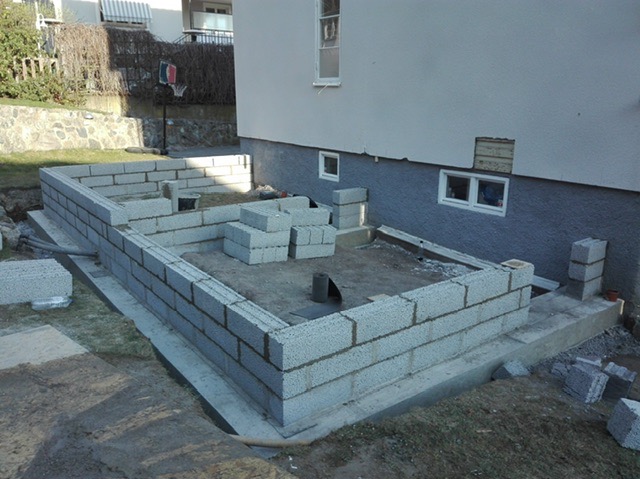 Wznoszenie ścian fundamentowych z bloczków betonowych. Widoczne ułożone bloczki, fundament i fragment budynku w tle. Prace murarskie w toku.