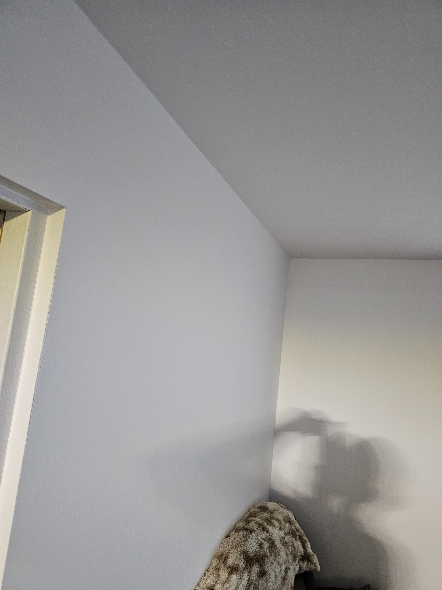 Świeżo pomalowany róg pokoju w odcieniach szarości, widoczny fragment futryny drzwi i cień na ścianie. Gładkie powierzchnie, minimalistyczny kadr.