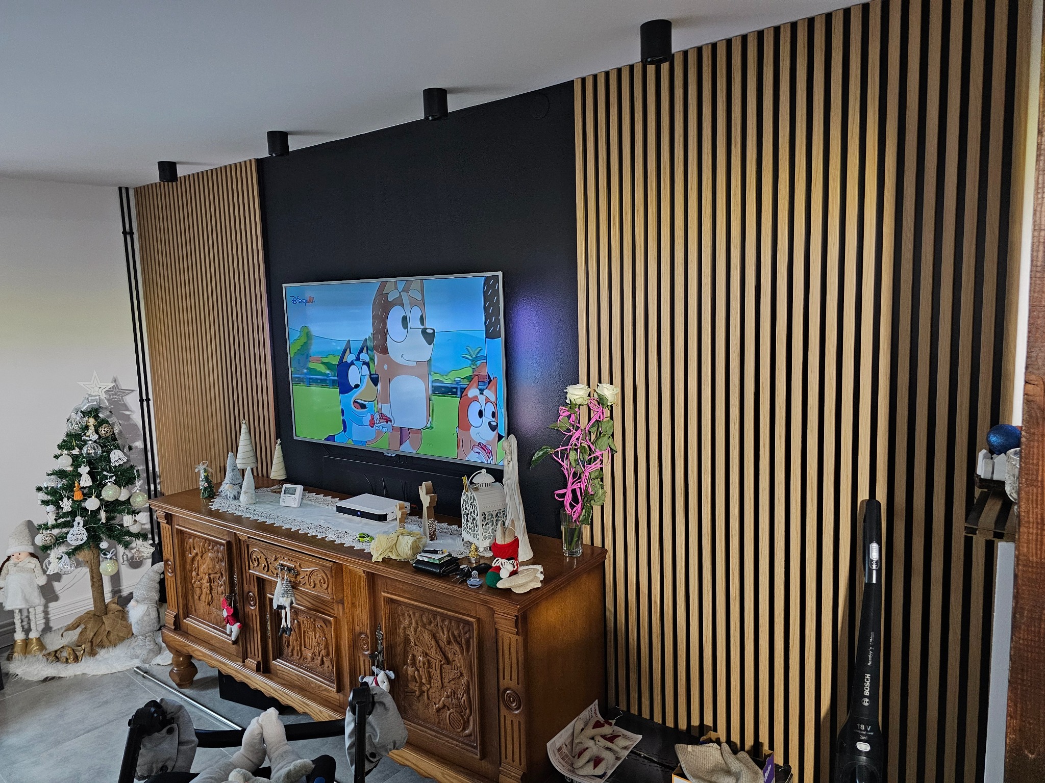 Salon z drewnianymi lamelami na ścianie za telewizorem, komoda z dekoracjami świątecznymi i kwiatami. Nowoczesny design wnętrza.