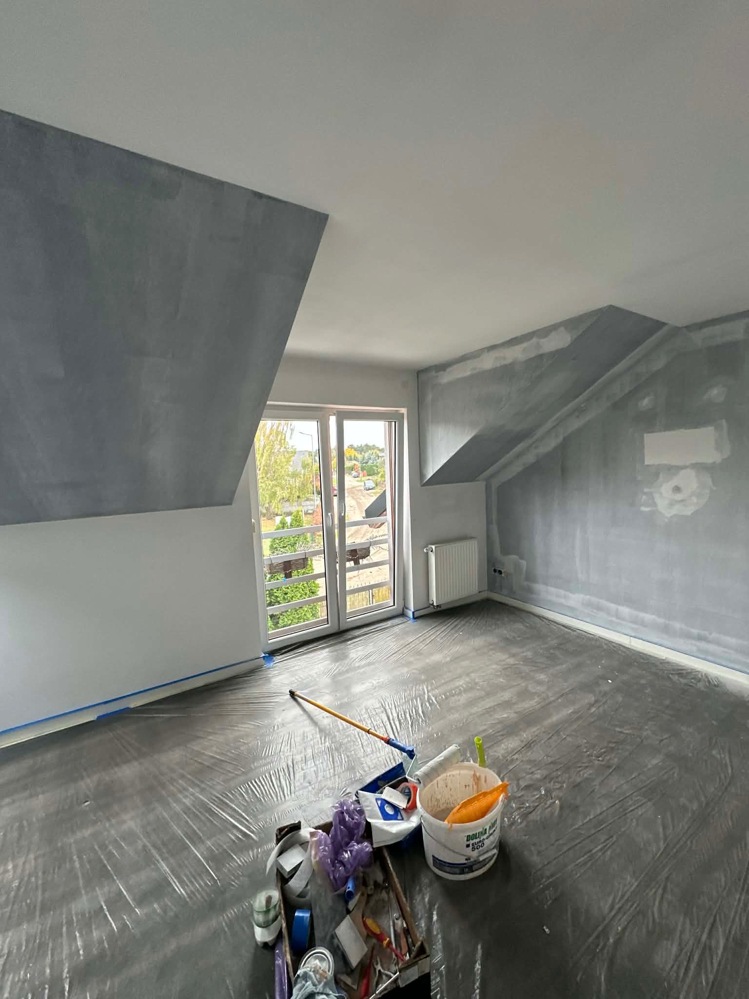 Pokój na poddaszu w trakcie prac wykończeniowych: ściany częściowo pomalowane na szaro, podłoga zabezpieczona folią, widoczne narzędzia malarskie i wiadro z farbą.