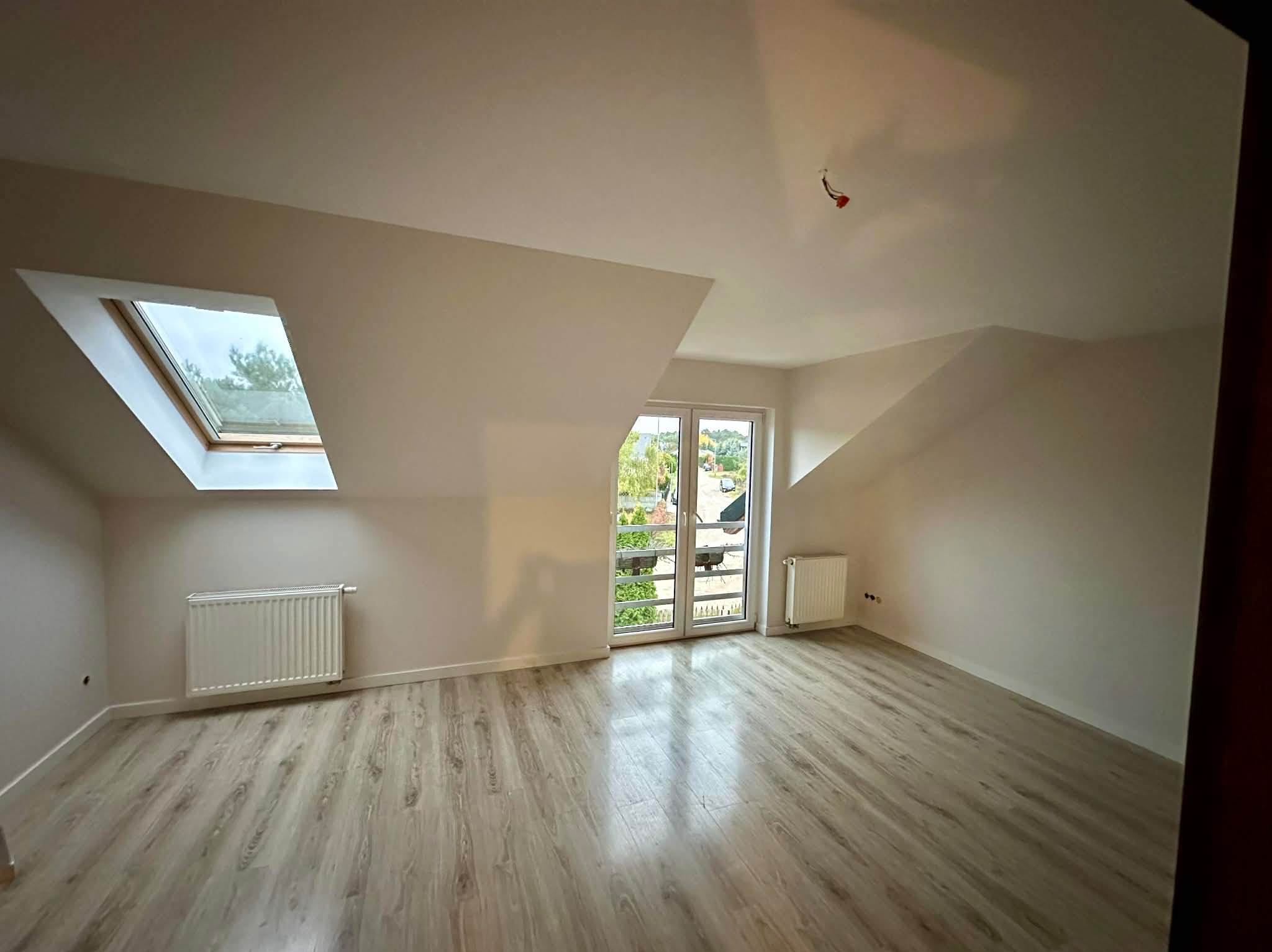 Jasne poddasze po malowaniu: skośne ściany, okno dachowe, parkiet, grzejniki. Widoczne świeżo pomalowane ściany w odcieniach bieli i beżu.