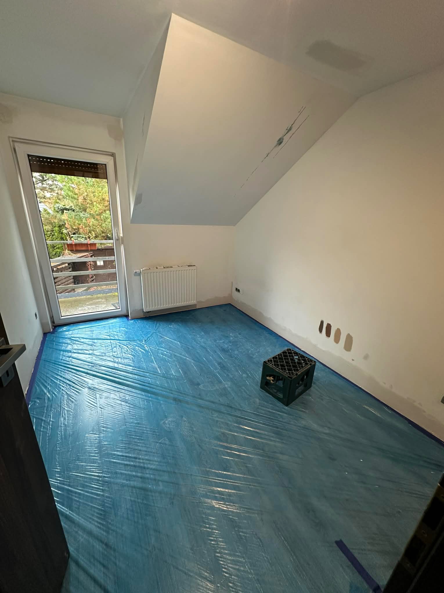Pokój w trakcie przygotowań do malowania: podłoga zabezpieczona folią, ściany z widocznymi śladami po szpachlowaniu, skrzynka na podłodze, okno balkonowe.