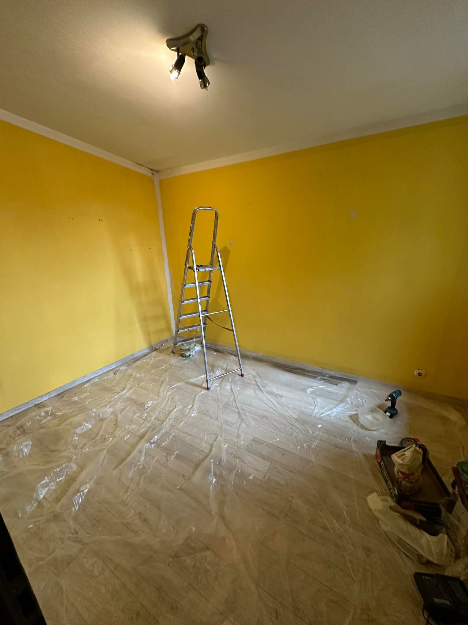 Pokój po malowaniu na żółto, zabezpieczona podłoga folią, drabina aluminiowa, widoczne narzędzia malarskie i wiertarka. Świeżo pomalowane ściany.