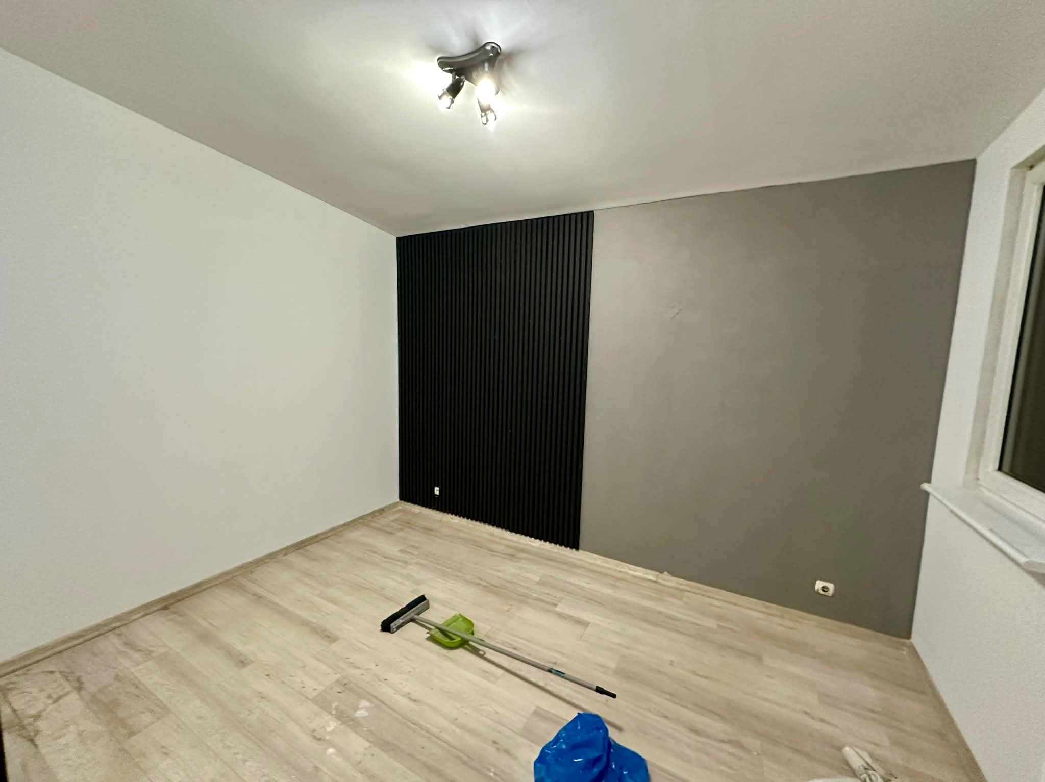 Pokój po remoncie: biała ściana kontrastuje z szarą i czarną ścianą z lamelami. Na podłodze leżą narzędzia malarskie, co sugeruje świeżo zakończone prace.