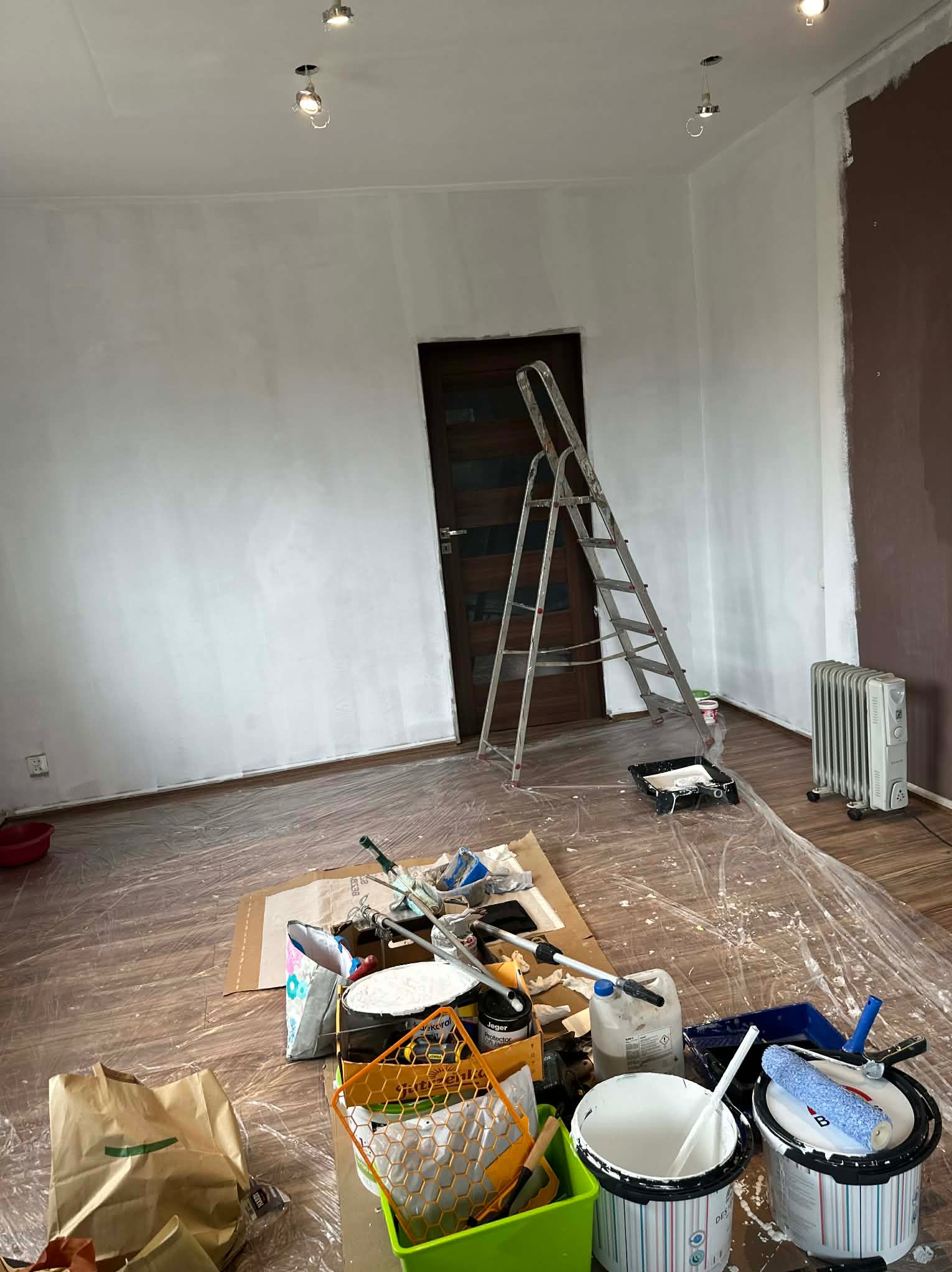 Pokój w trakcie odświeżania: częściowo pomalowane ściany na biało, drabina oparta o ścianę, liczne narzędzia malarskie rozłożone na folii ochronnej na podłodze.