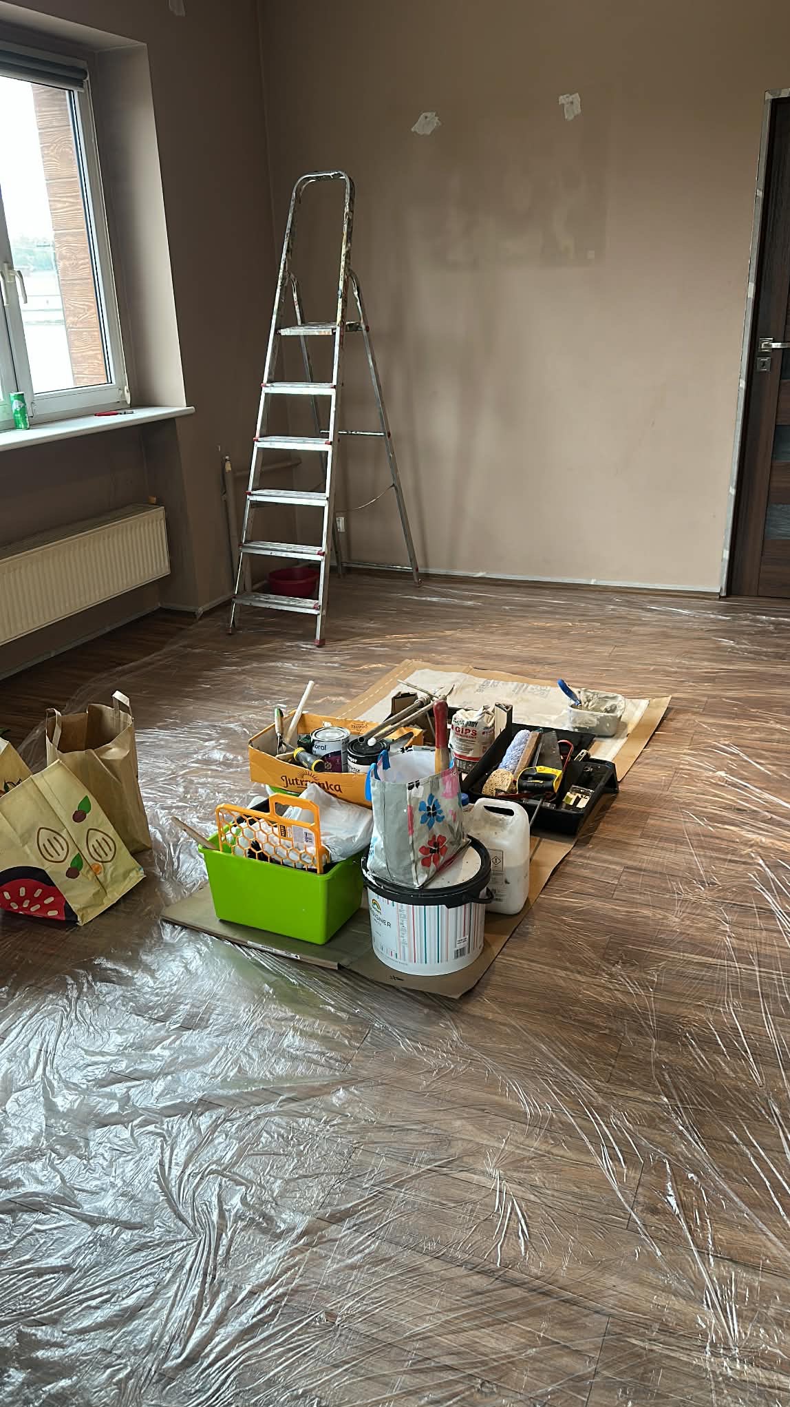 Pokój przygotowany do malowania: ściany po szpachlowaniu, zabezpieczona podłoga folią, drabina i akcesoria malarskie w centrum kadru. Widok na okno i grzejnik.
